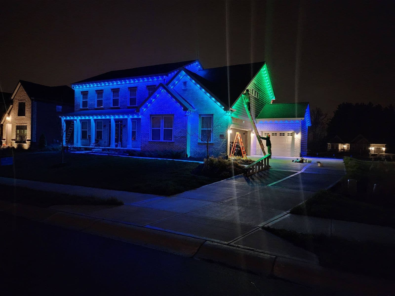 Worker on ladder installing blue and green LED lights on house roofline at night
