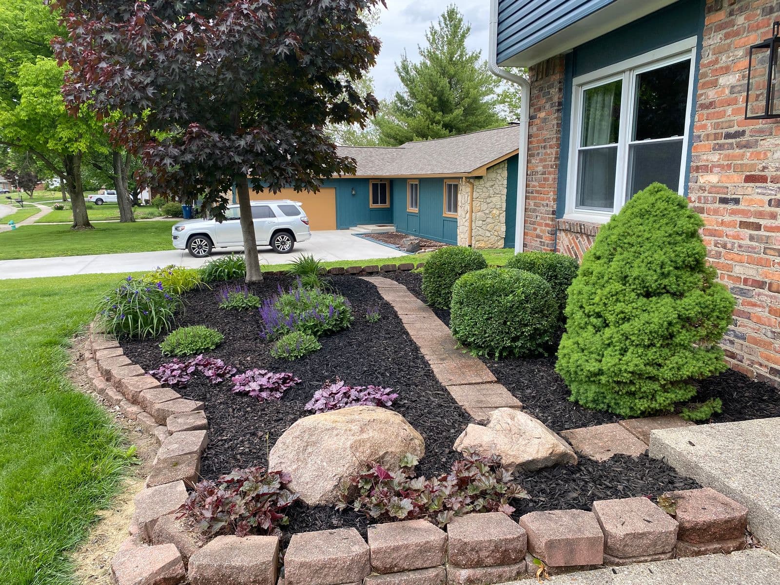 Front yard flower bed with black mulch, boulders, trimmed shrubs, and brick edging